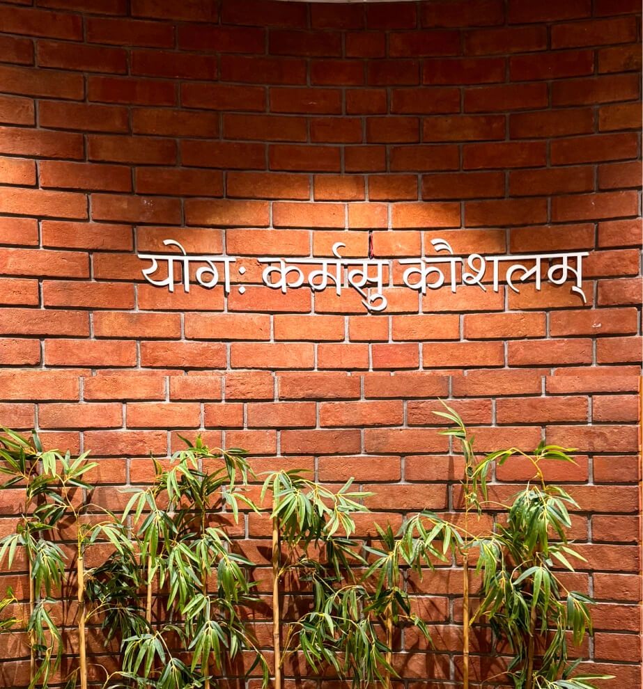 A red brick wall in Aakash’s Ahmedabad office, with the Sanskrit line योगः कर्मसु कौशलम् mounted in white letters above a row of bamboo.